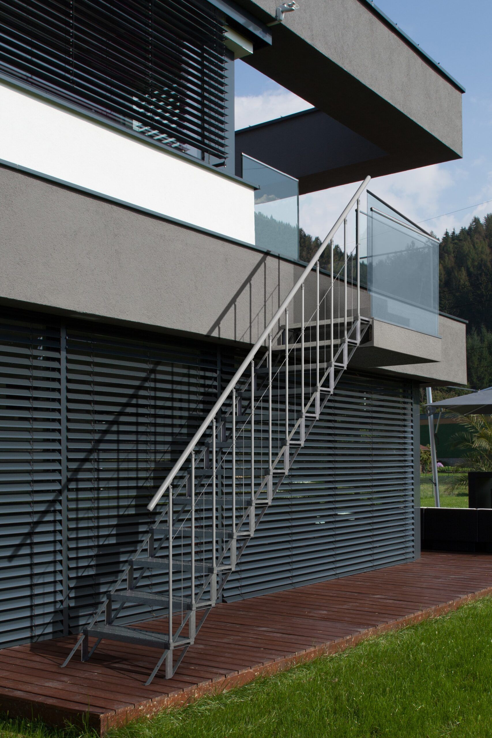 Outdoor staircase Sun Step made of galvanized steel on a modern house facade. The staircase has 12 separate grid steps, a railing with vertical bars on both sides, and a stainless steel handrail. The stairs connect the terrace to the garden.