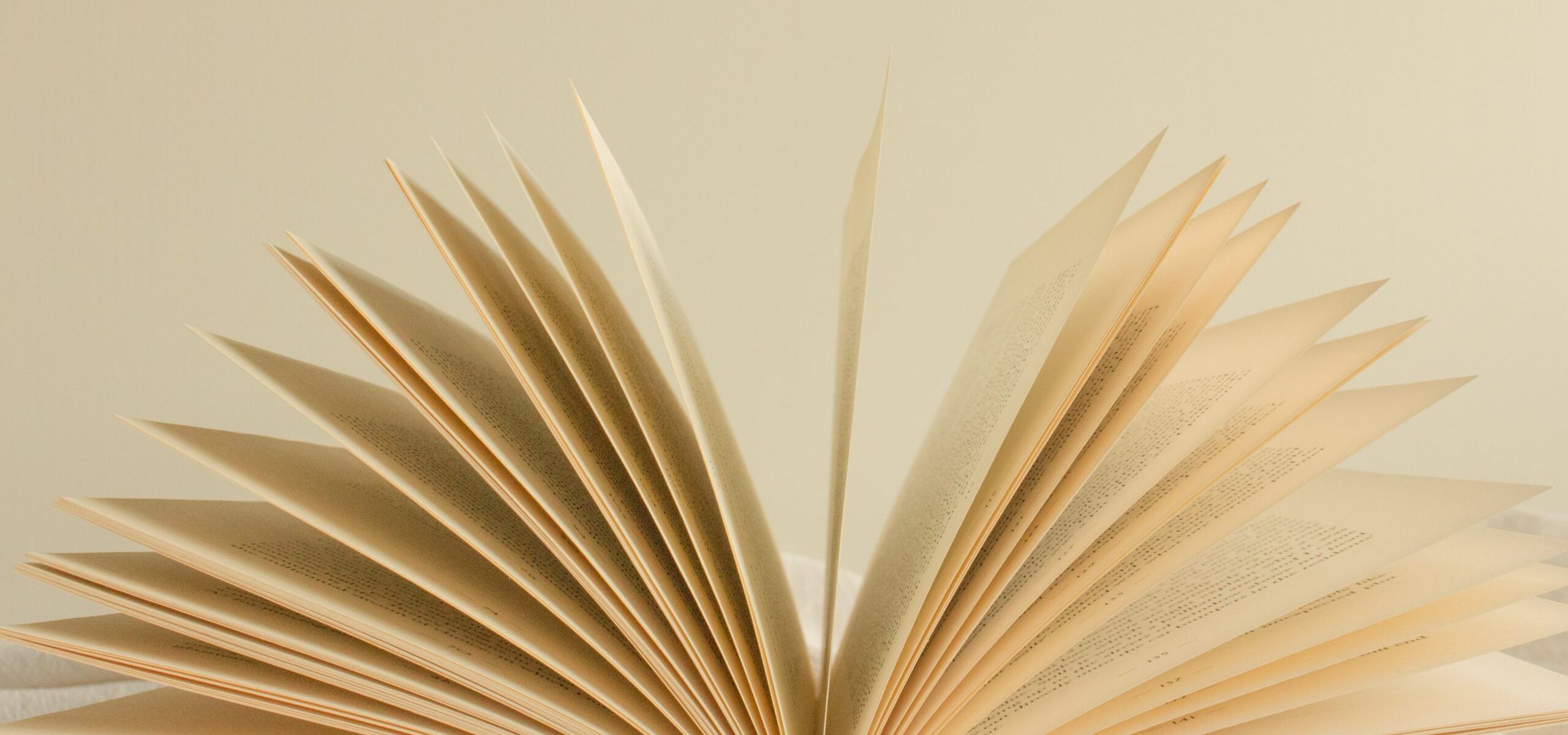 An open book with pages fanned out, revealing lines of printed text on yellowed paper. The book is placed on a soft, neutral-colored surface.