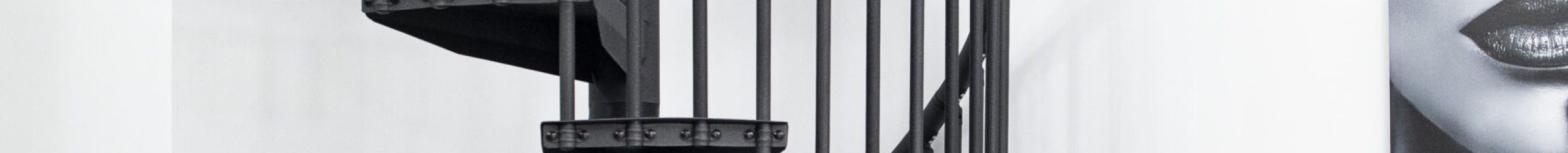 Spiral staircase Rondo Color in anthracite in a bright, modern interior. The steps and vertical bar railing are also in anthracite. The space-saving staircase connects the lower and upper floor and stands against a white wall.