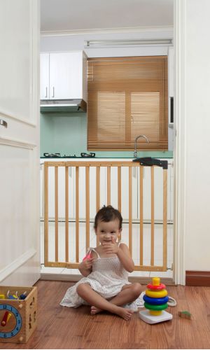 A toddler plays with colorful toys on the floor in front of the mounted "Annika" wooden child safety gate. The gate is installed between two walls, separating the play area from the kitchen behind.