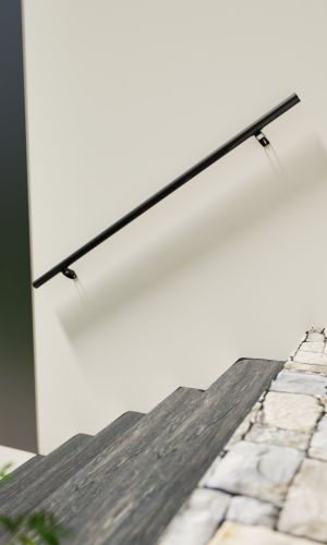 Side view of the Sirius handrail in anthracite, mounted on a light-colored wall along a modern staircase with dark steps and contrasting white stringers. The handrail is sleek, round, and supported by minimalist wall brackets.