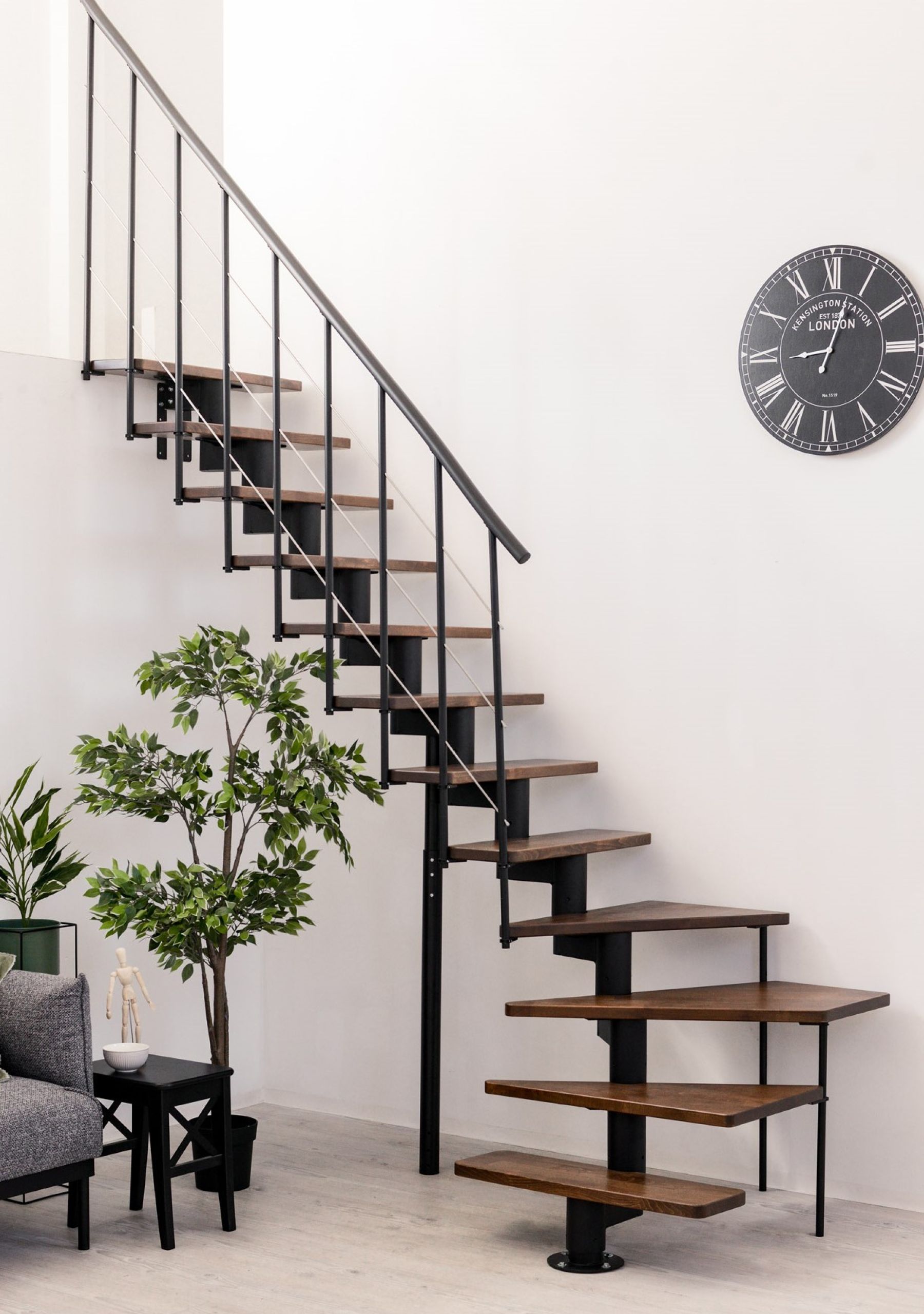 The image shows a space-saving staircase, featuring a stylish design with floating wooden steps supported by a sleek black metal frame. The staircase leads to an upper level in a modern living space, complemented by greenery and a clock on the wall. This type of staircase is typically used to maximize space in smaller areas while still maintaining an aesthetic appeal.