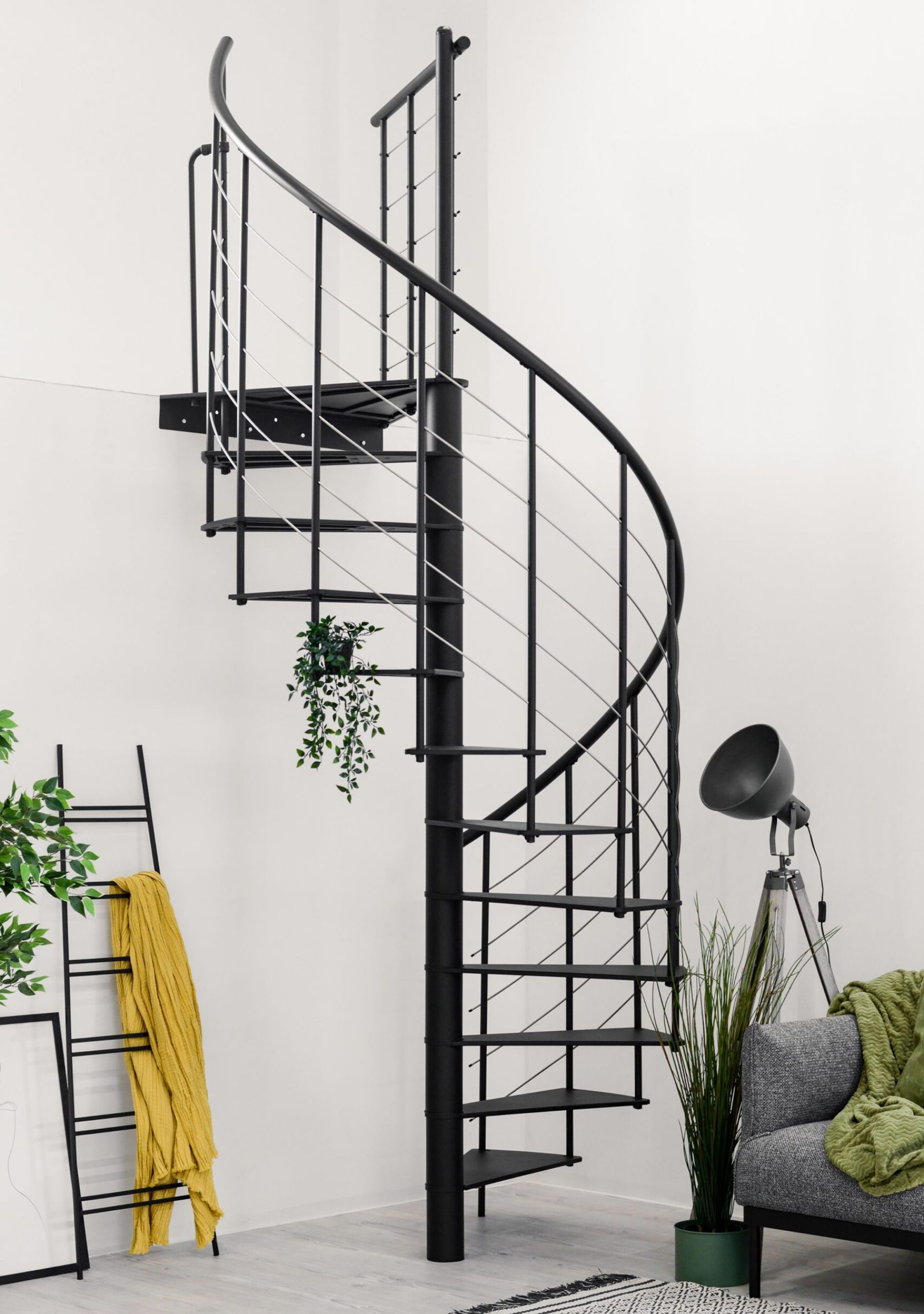 The image shows a modern spiral staircase, which features a sleek black metal frame. It is located in a contemporary indoor setting, with light-colored walls, minimalistic furniture, and decorative plants surrounding the staircase.
