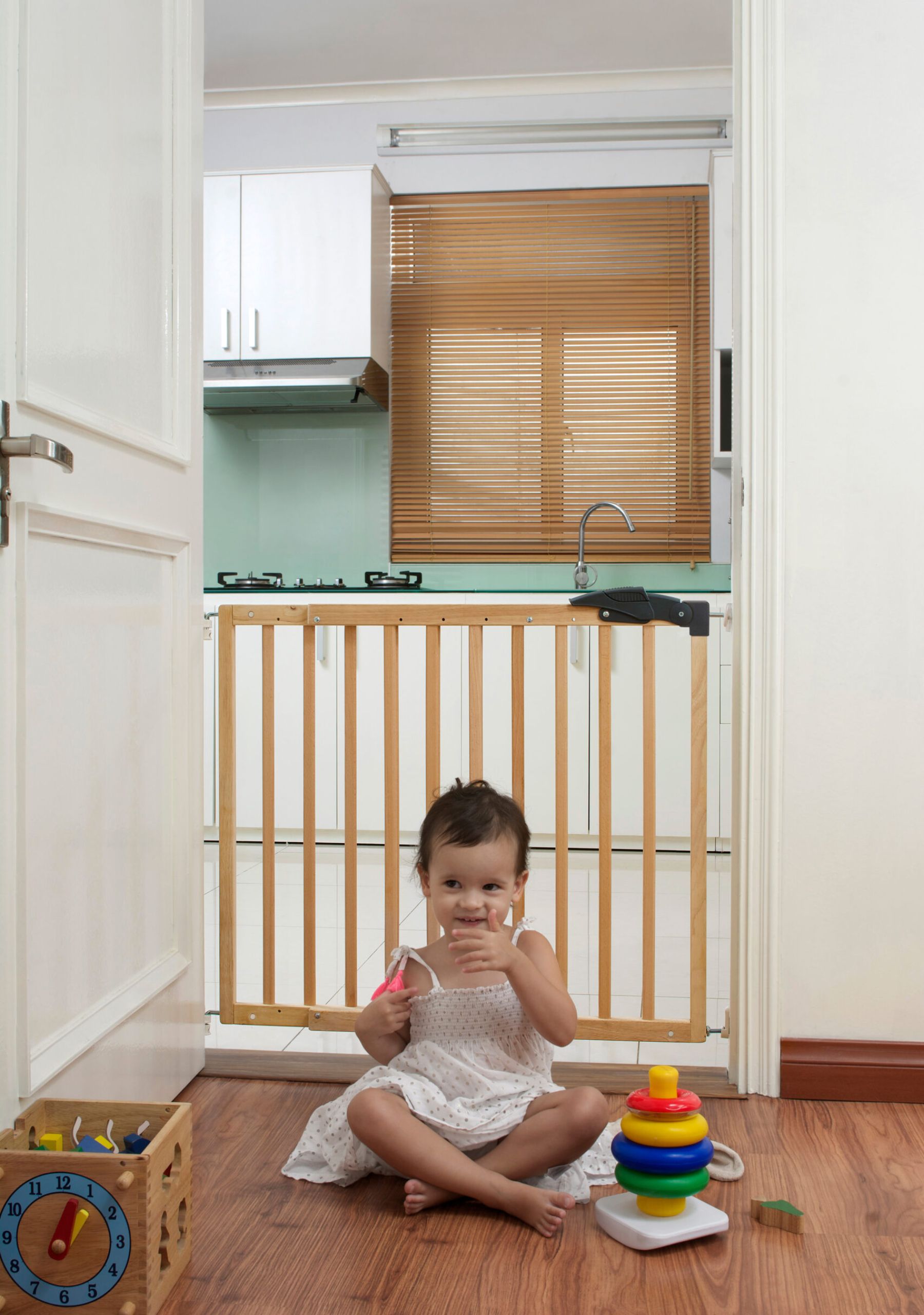 A small girl sits on a wooden floor in front of the closed “Erik” child safety gate, which is installed in a doorway between the living room and kitchen. Colorful toys are scattered around her in the foreground.