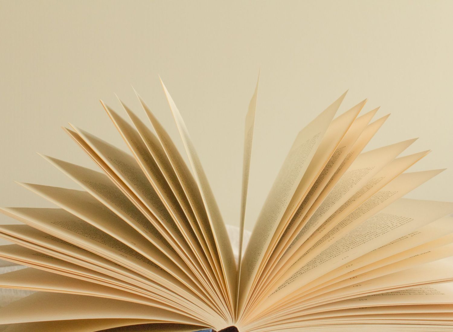 An open book with pages fanned out, revealing lines of printed text on yellowed paper. The book is placed on a soft, neutral-colored surface.