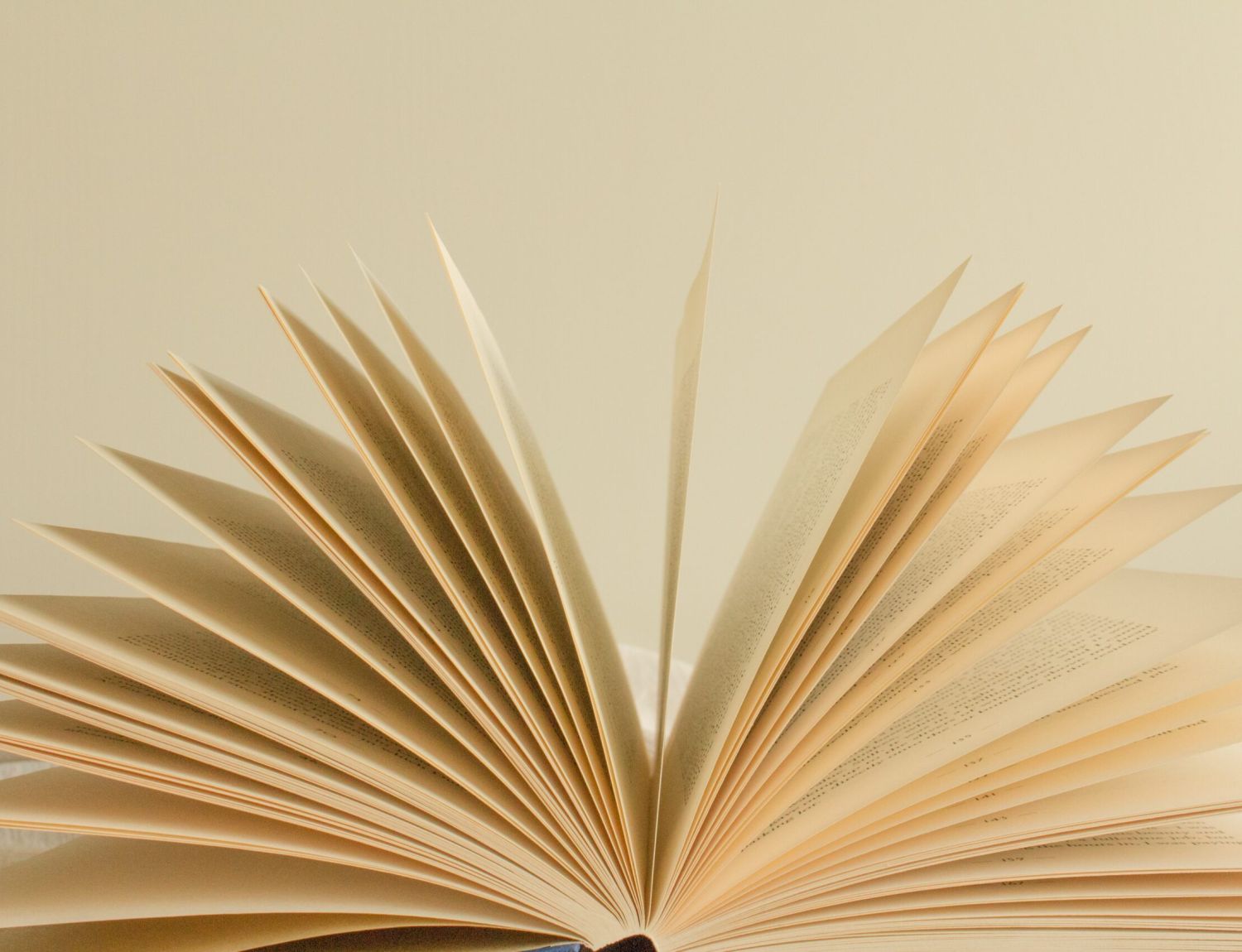 An open book with pages fanned out, revealing lines of printed text on yellowed paper. The book is placed on a soft, neutral-colored surface.