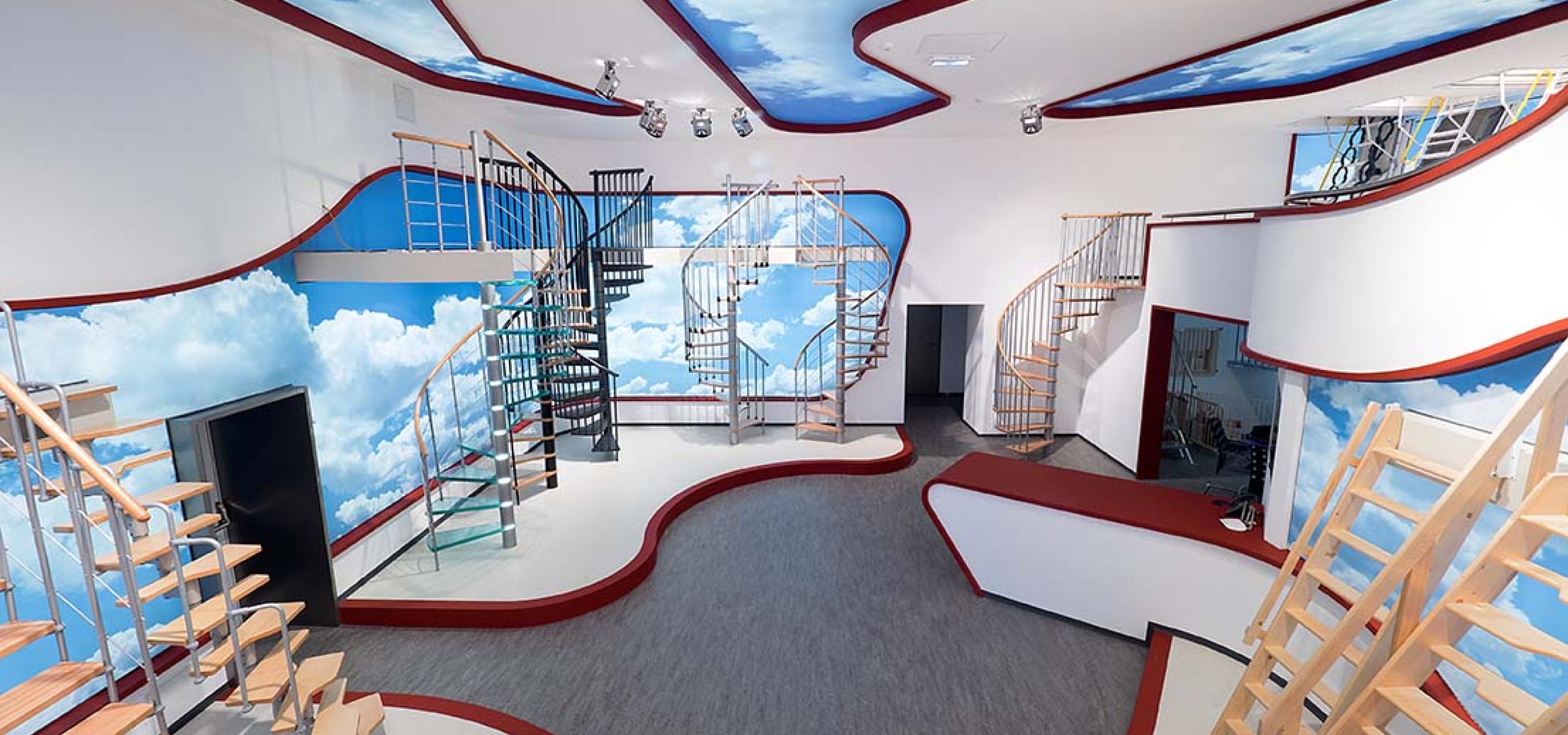 Wide-angle view of the modern Minka showroom featuring various staircase models. The sky-themed walls and ceiling create an open, inspiring atmosphere.