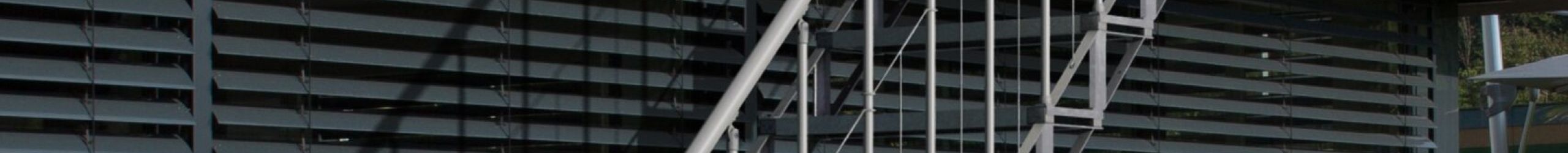 Outdoor staircase Sun Step made of galvanized steel on a modern house facade. The staircase has 12 separate grid steps, a railing with vertical bars on both sides, and a stainless steel handrail. The stairs connect the terrace to the garden.