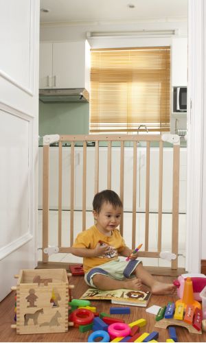 A toddler plays with colorful toys on the floor in front of the mounted "Annika" wooden child safety gate. The gate is installed between two walls, separating the play area from the kitchen behind.