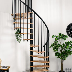 The Berlin spiral staircase with beech steps and black banisters is mounted on a gallery. The room is decorated with plants, a black wall clock, a black side table and a patterned carpet.