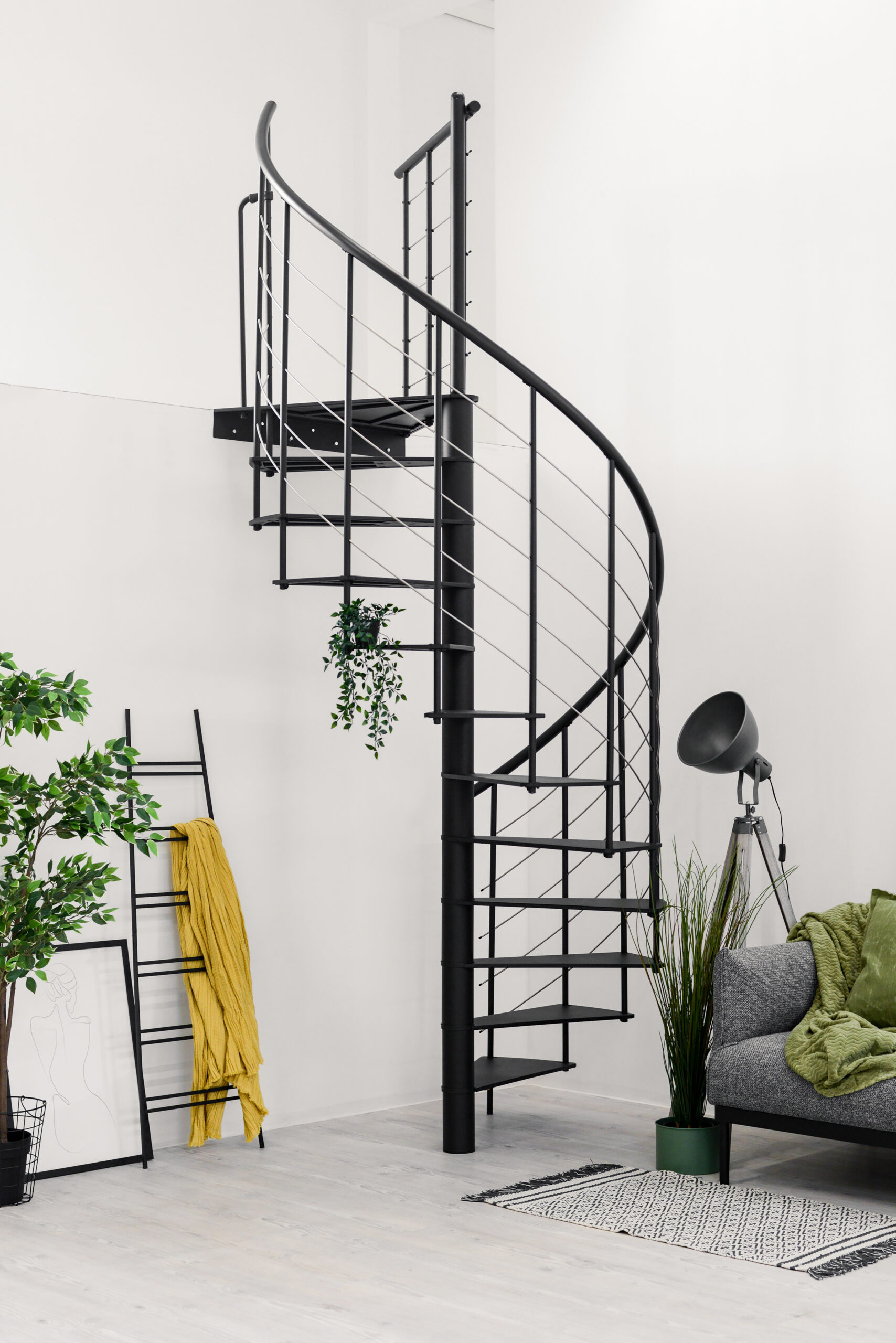 Modern “Milano” spiral staircase made of black metal. The staircase features open metal steps, a railing with vertical rods, horizontal base bars (underguards), and a black PVC handrail.