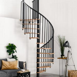 Complete shot of the Spiral Smart spiral staircase with beech steps and black banister, installed in a white room. On the left is a grey sofa with white and brown cushions and a black side table with decorations. On the right is a grey floor lamp, a houseplant and a raffia basket.