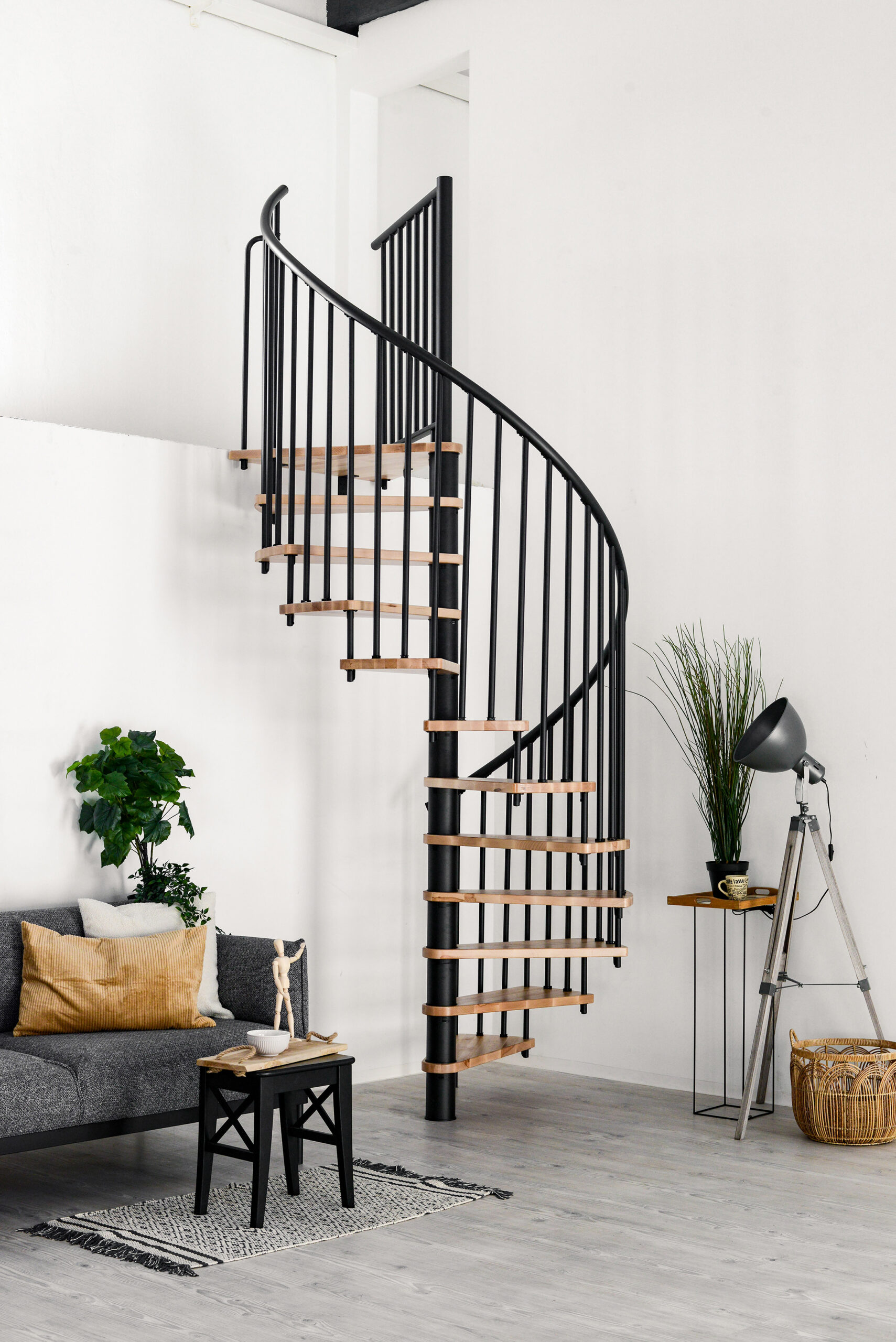 Complete shot of the Spiral Smart spiral staircase with beech steps and black banister, installed in a white room. On the left is a grey sofa with white and brown cushions and a black side table with decorations. On the right is a grey floor lamp, a houseplant and a raffia basket.