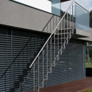 Outdoor staircase Sun Step made of galvanized steel on a modern house facade. The staircase has 12 separate grid steps, a railing with vertical bars on both sides, and a stainless steel handrail. The stairs connect the terrace to the garden.