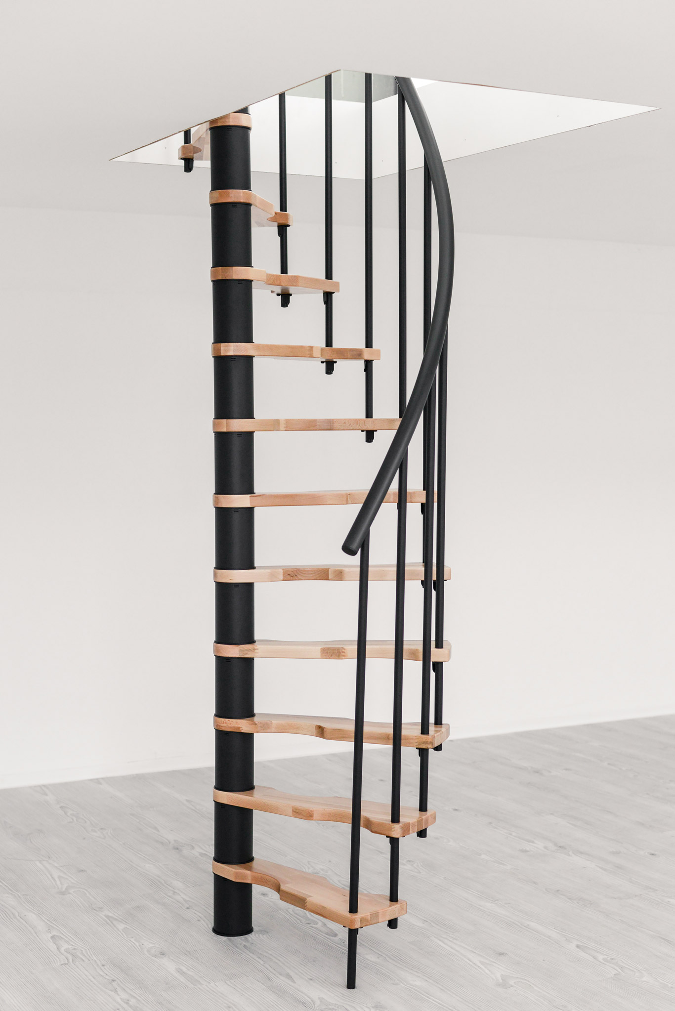 The spiral staircase Suono Smart with black PVC handrail, black railing rods, and light wooden steps impresses with its modern design and space-saving construction.