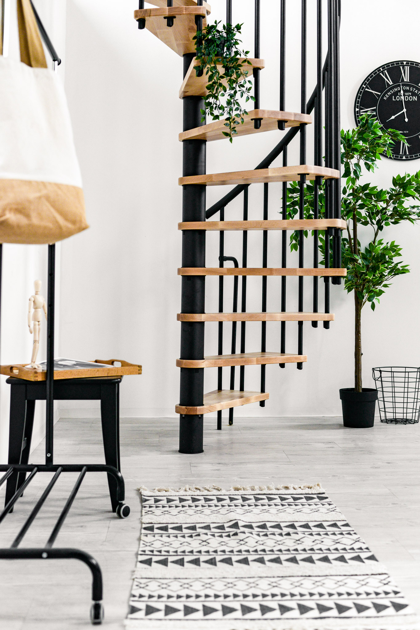 Spiral staircase Berlin in a cosy room with a patterned carpet, houseplants, wall clock and black decorations. The beech steps with the steel banister revolve around the black metal spindle of the staircase