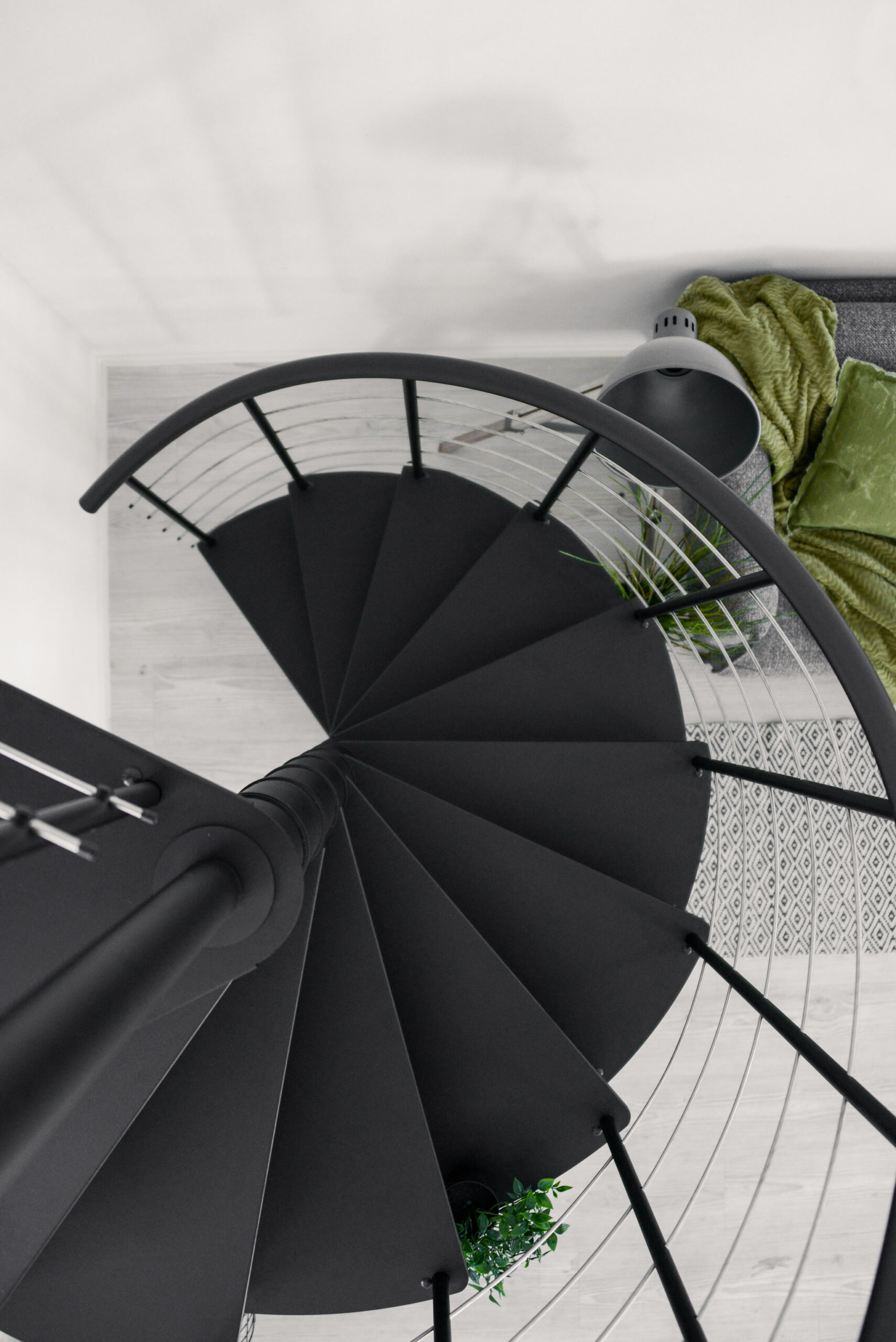 Top-down view of the black “Milano” spiral staircase with triangular metal steps and a curved PVC handrail. The railing features vertical metal rods with horizontal base bars.