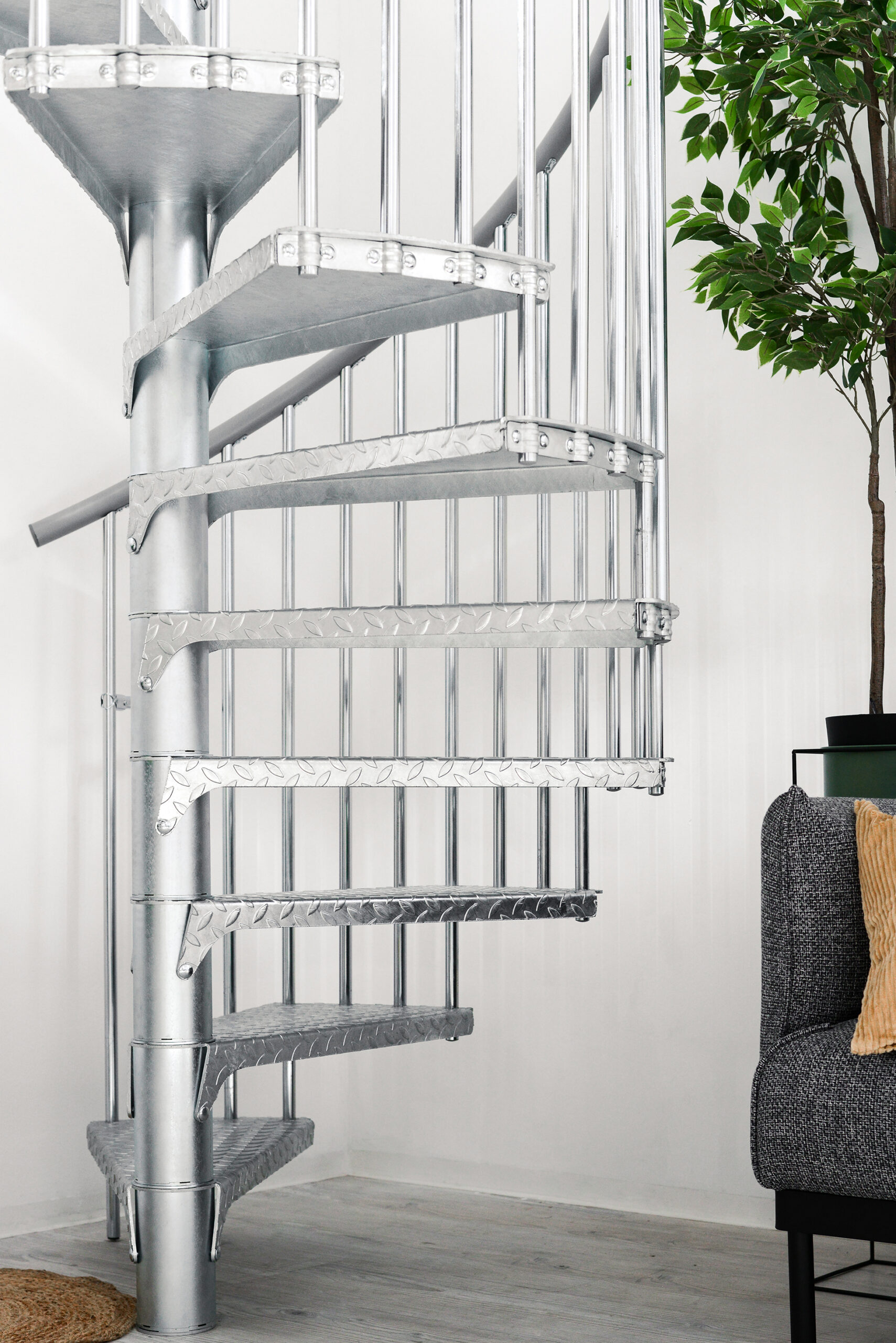 Close-up of the outdoor spiral staircase Rondo Zink Smart. Visible are galvanized metal steps with a tread plate structure and round railing bars. On the right side a grey sofa and a green plant.