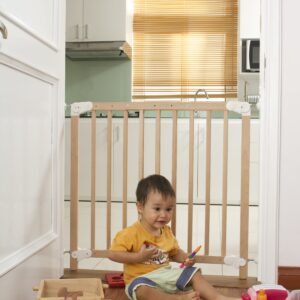 A toddler plays with colorful toys on the floor in front of the mounted "Annika" wooden child safety gate. The gate is installed between two walls, separating the play area from the kitchen behind.