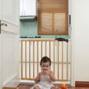 A small girl sits on a wooden floor in front of the closed “Erik” child safety gate, which is installed in a doorway between the living room and kitchen. Colorful toys are scattered around her in the foreground.