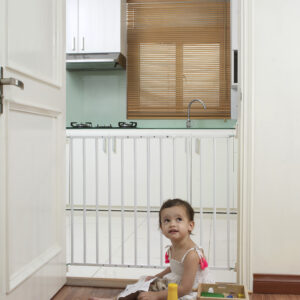 A small girl is playing with toys on the floor in front of the “Felix” child safety gate, which is mounted in a doorway. The gate is made of white metal bars and separates the living area from the kitchen.