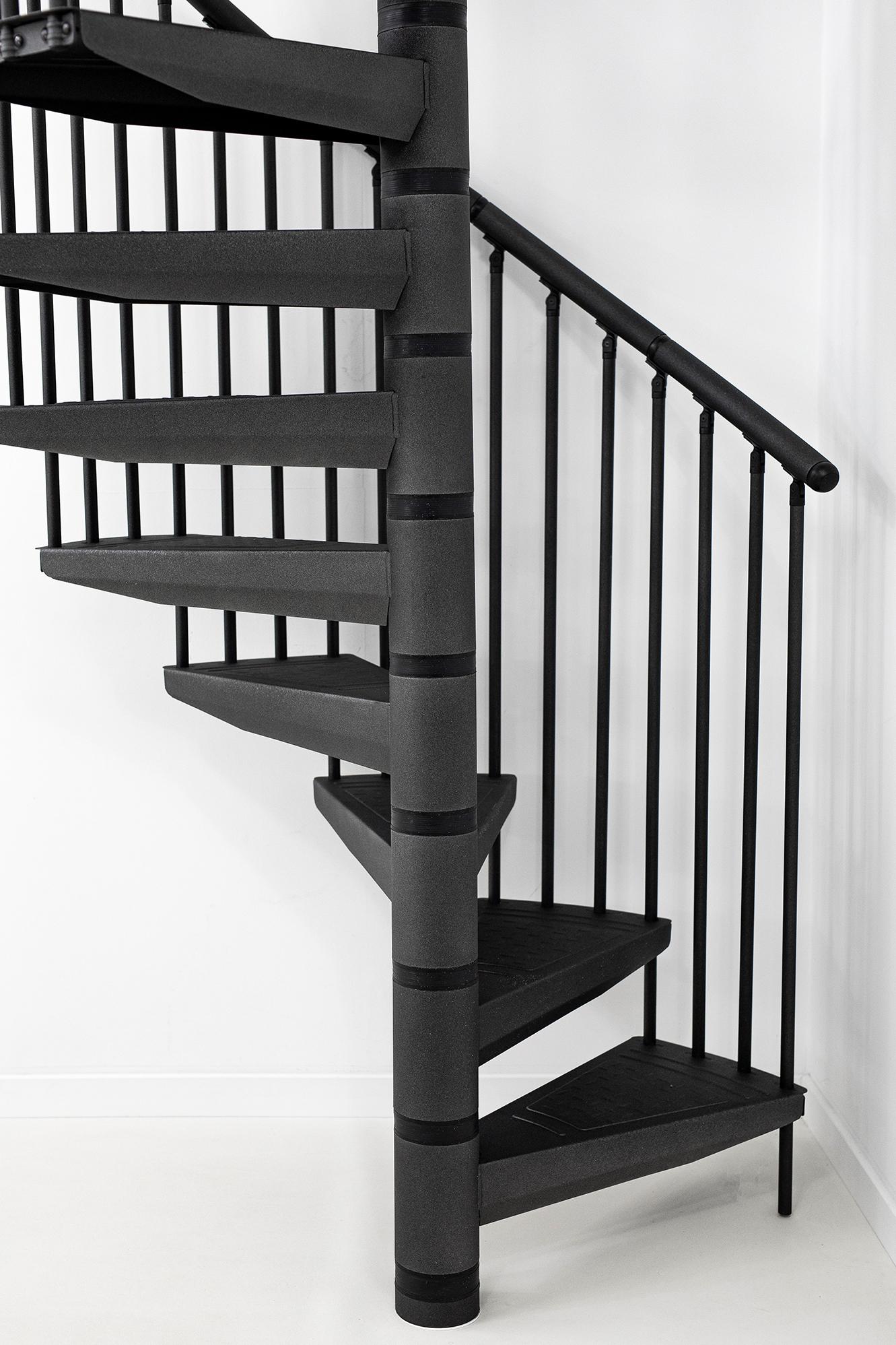Detail view of the spiral staircase Rondo Color in anthracite. The image shows the lower steps, the central column and the vertical bar railing. The staircase is made of powder-coated metal and stands against a white wall.