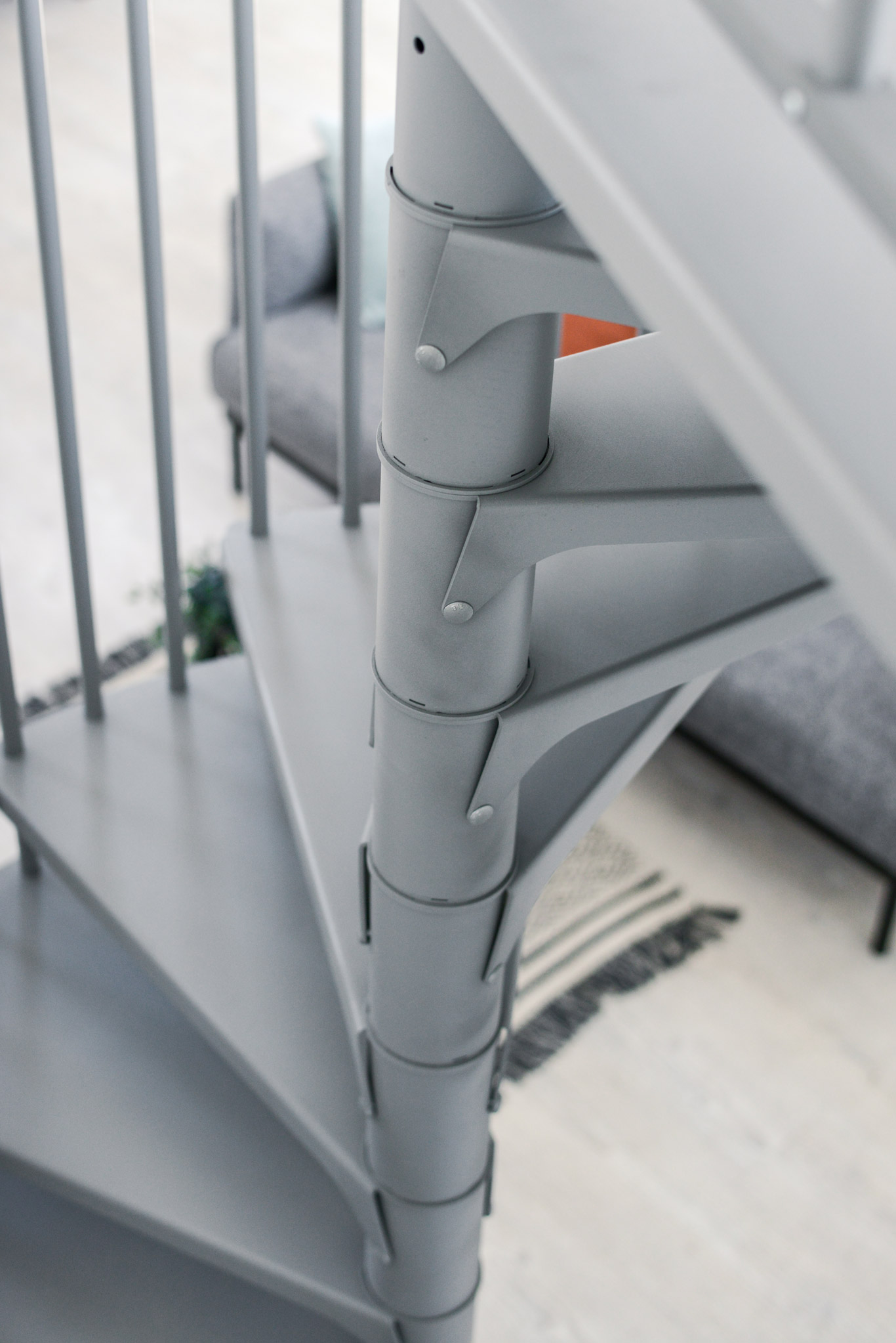 Close-up view of the spiral staircase Rondo Smart in grey. The central metal column with connected steps is visible. In the background, a grey sofa and a rug on light wooden flooring.