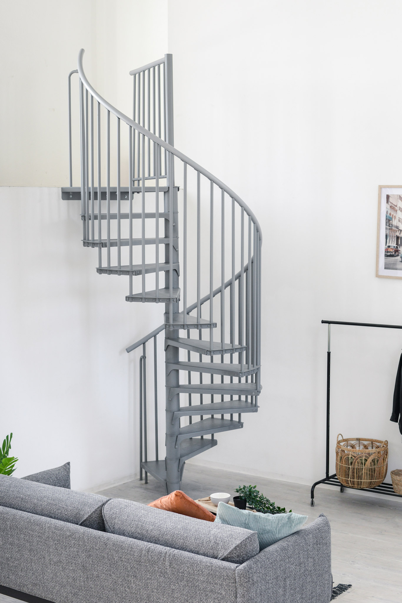 Grey metal spiral staircase Rondo Smart with round steps and vertical railing rods. The space-saving staircase winds upwards in a modern living area with sofa, plants and a clothes rack.