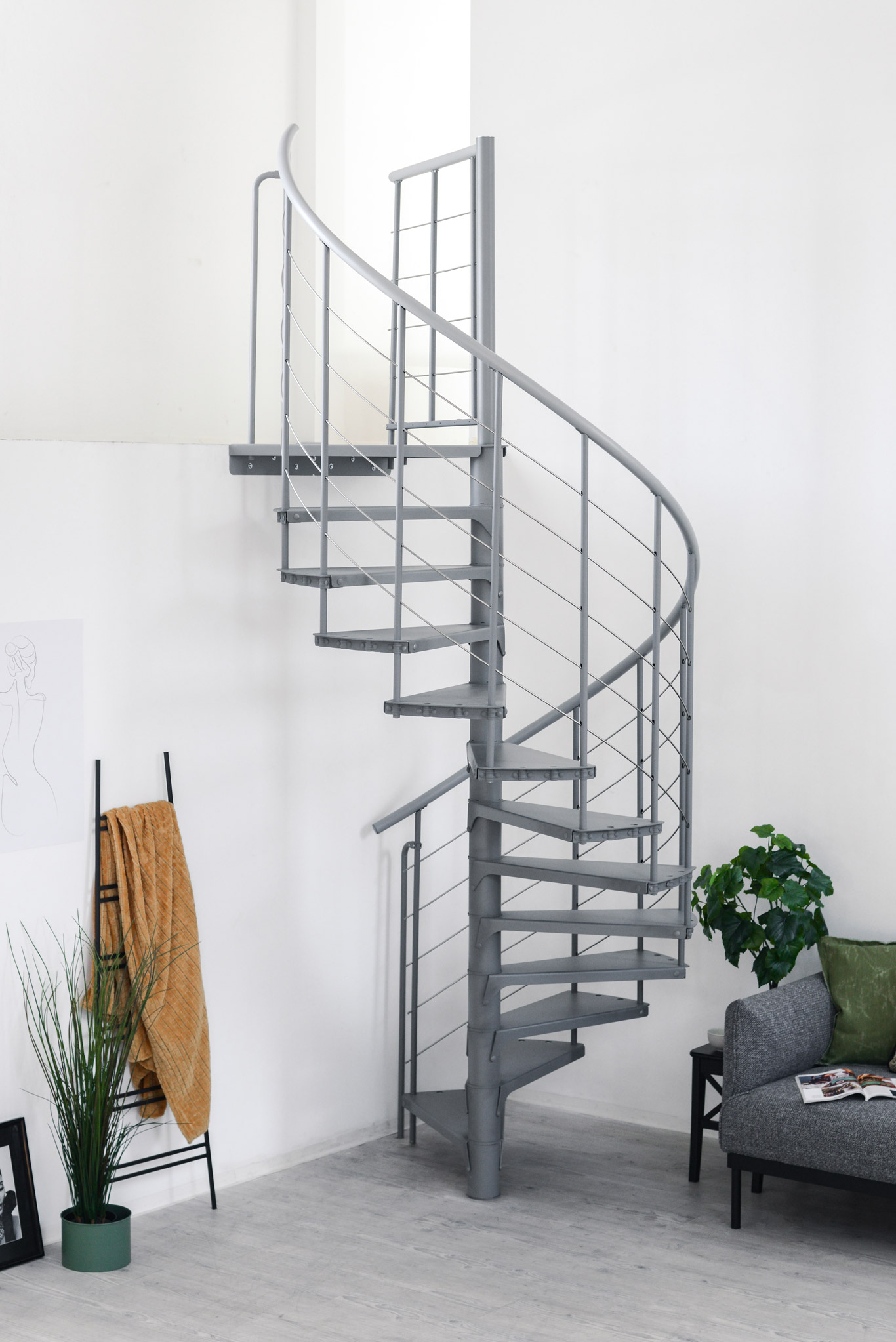 Spiral staircase Spino Smart in grey metal in a modern living space. The winding stairs lead upwards, the railing consists of vertical rods and stainless steel cables. In the background are a sofa, plants, and decorations.