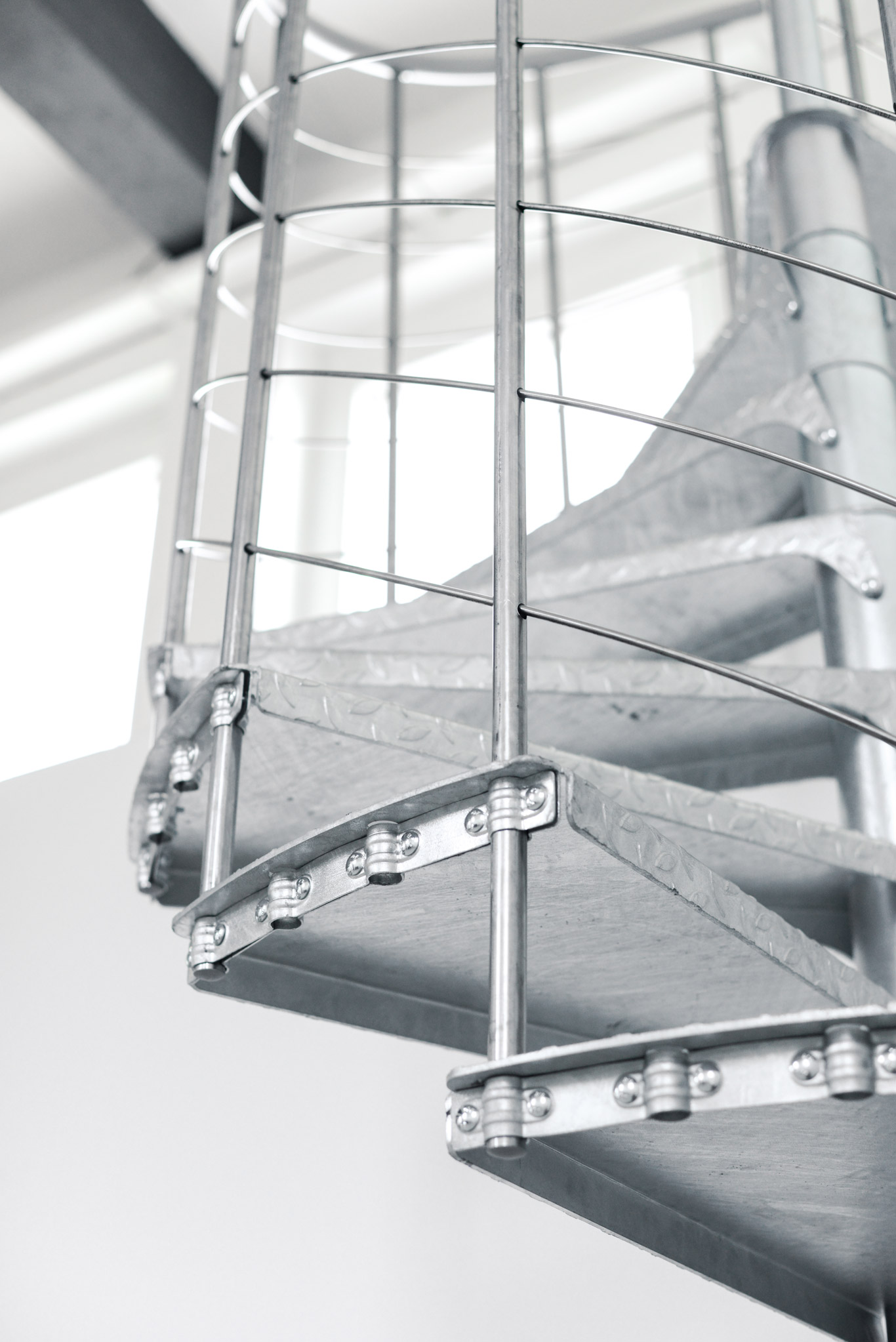 Close-up of the Spino Zink Smart spiral staircase featuring galvanized metal steps and a railing with vertical and horizontal bars. The sturdy construction ensures safety and accessibility in both indoor and outdoor environments.