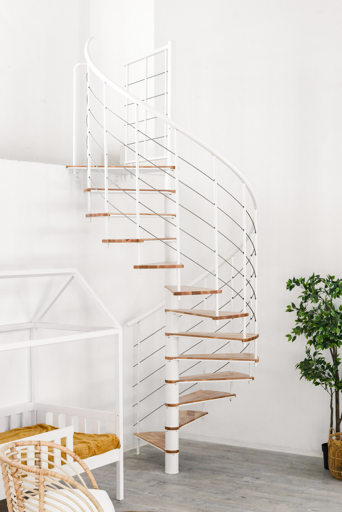 Fusion spiral staircase with beech steps and white metal banister built into a bright room with plants, cot and chair.
