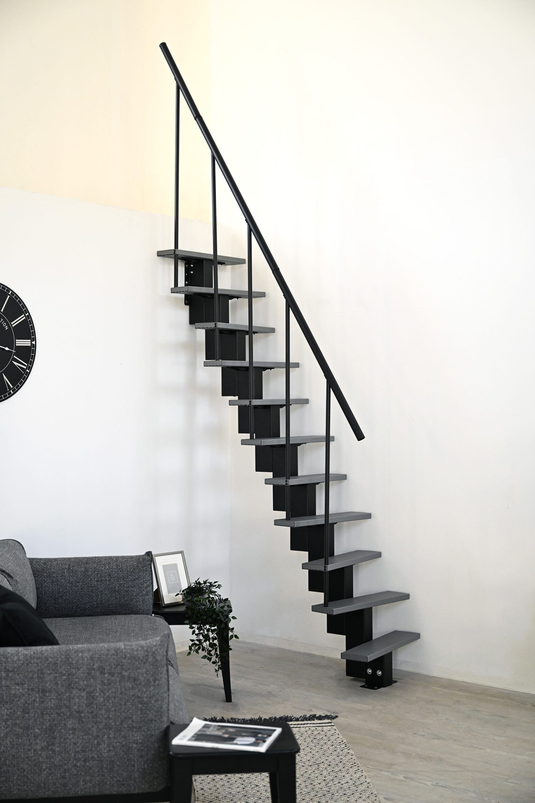 Modern space-saving staircase with alternating offset steps in grey and black metal structure. The staircase is straight, narrow and rises steeply up a white wall. A simple black banister provides support.
