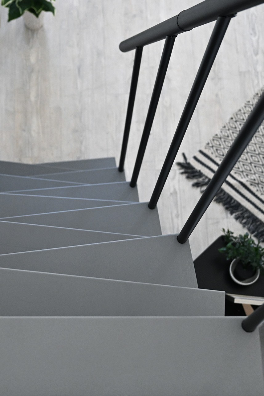 Detailed view of a straight metal staircase with irregularly shaped grey steps. The staircase has a black handrail with vertical round bars.