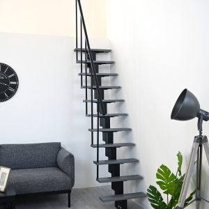Room-saving staircase with staggered grey treads and black central stringer in a modern living room with a light wooden floor. The staircase is fitted with a black handrail on one side. On the left is a grey sofa, on the right a black floor lamp next to a green plant in a wire basket.
