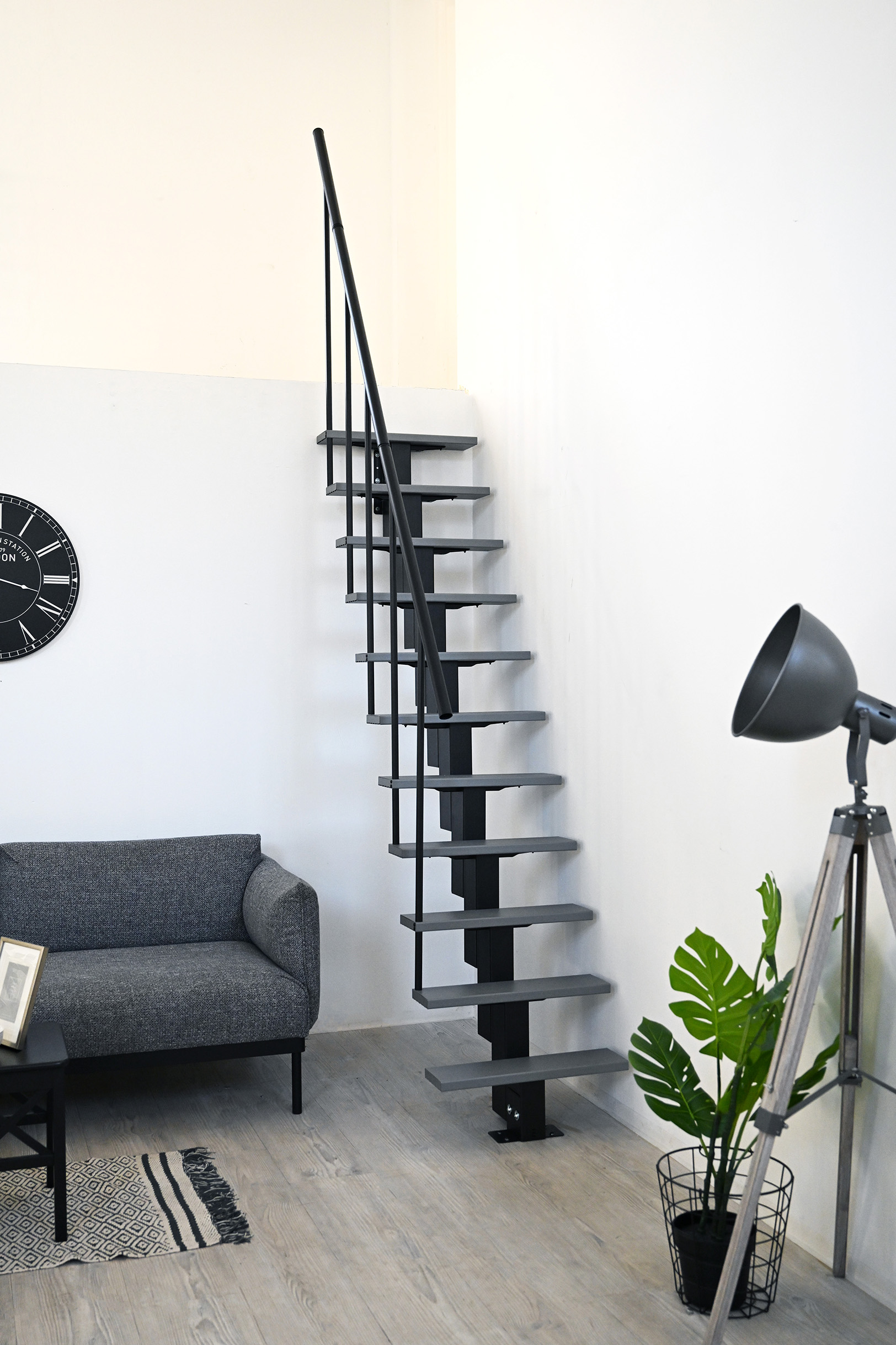 Room-saving staircase with staggered grey treads and black central stringer in a modern living room with a light wooden floor. The staircase is fitted with a black handrail on one side. On the left is a grey sofa, on the right a black floor lamp next to a green plant in a wire basket.