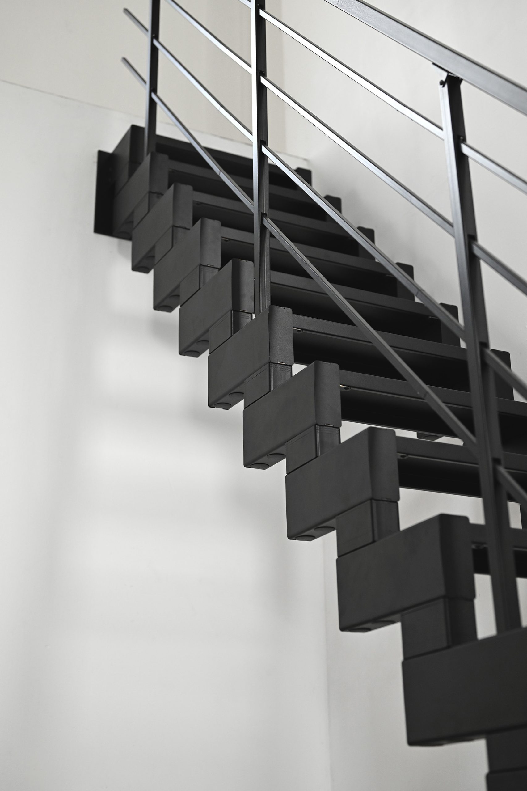 Detailed view of the black Modulario Mini space-saving staircase from below. The staggered metal treads and the modern banister with horizontal struts are visible. The staircase is mounted on a white wall.