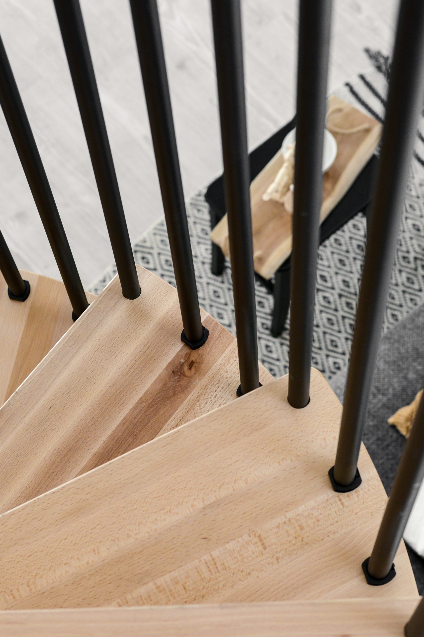 Close-up of the beech steps of the spiral staircase Spiral Effect from above. The black steel bars of the banister are attached to the outside of the steps. Behind them you can see a patterned carpet with a black table.