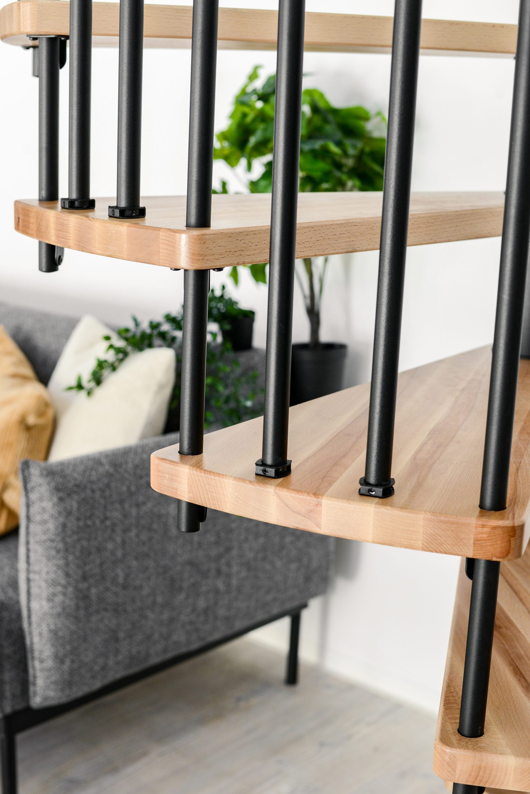 Close-up of the spiral staircase Spiral Effect with beech steps and black balusters from the side. In the background is a grey couch with plants and cushions.