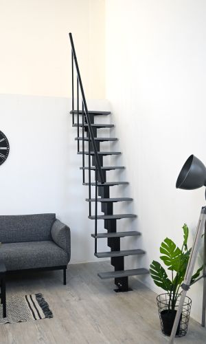Room-saving staircase with staggered grey treads and black central stringer in a modern living room with a light wooden floor. The staircase is fitted with a black handrail on one side. On the left is a grey sofa, on the right a black floor lamp next to a green plant in a wire basket.