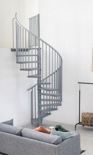 Grey metal spiral staircase Rondo Smart with round steps and vertical railing rods. The space-saving staircase winds upwards in a modern living area with sofa, plants and a clothes rack.