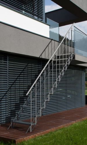 Outdoor staircase Sun Step made of galvanized steel on a modern house facade. The staircase has 12 separate grid steps, a railing with vertical bars on both sides, and a stainless steel handrail. The stairs connect the terrace to the garden.