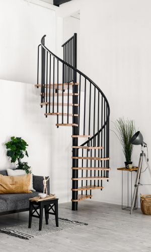 Complete shot of the Spiral Smart spiral staircase with beech steps and black banister, installed in a white room. On the left is a grey sofa with white and brown cushions and a black side table with decorations. On the right is a grey floor lamp, a houseplant and a raffia basket.