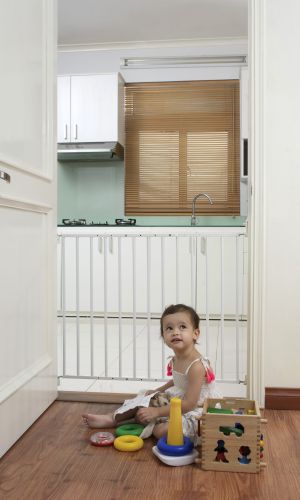 A small girl is playing with toys on the floor in front of the “Felix” child safety gate, which is mounted in a doorway. The gate is made of white metal bars and separates the living area from the kitchen.