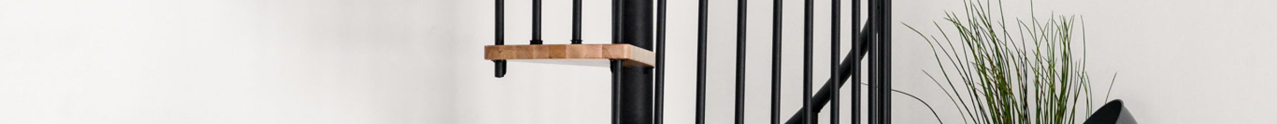 Gesamtaufnahme der Spindeltreppe Spiral Smart mit Buchenstufen und schwarzem Geländer, montiert in einem weißen Raum. Links steht ein graues Sofa mit weiß-braunen Kissen und ein schwarzer Beistelltisch mit Deko. Rechts steht eine graue Stehlampe, eine Zimmerpflanze und ein Bastkorb.