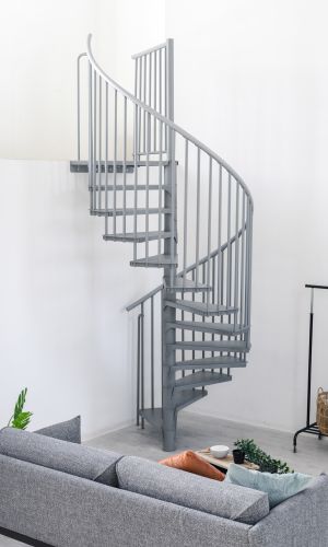 Graue Spindeltreppe Rondo Smart aus Metall mit runden Stufen und senkrechten Geländerstäben. Die platzsparende Treppe führt spiralförmig nach oben und steht in einem modernen Wohnraum mit Sofa, Pflanzen und Kleiderständer.