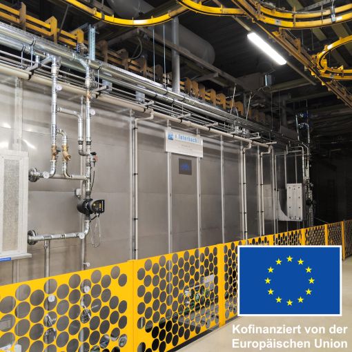 Foto einer modernen Industrieanlage mit Rohrleitungen und Schutzgittern. Rechts unten ist die EU-Flagge mit dem Hinweis „Kofinanziert von der Europäischen Union“ eingeblendet.