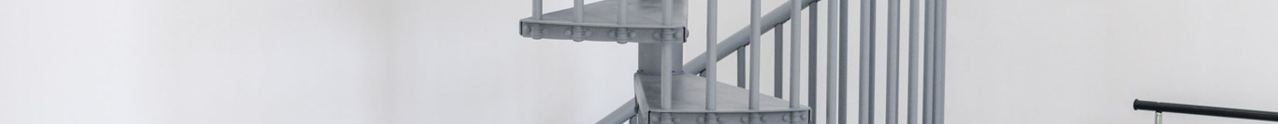 Graue Spindeltreppe Rondo Smart aus Metall mit runden Stufen und senkrechten Geländerstäben. Die platzsparende Treppe führt spiralförmig nach oben und steht in einem modernen Wohnraum mit Sofa, Pflanzen und Kleiderständer.