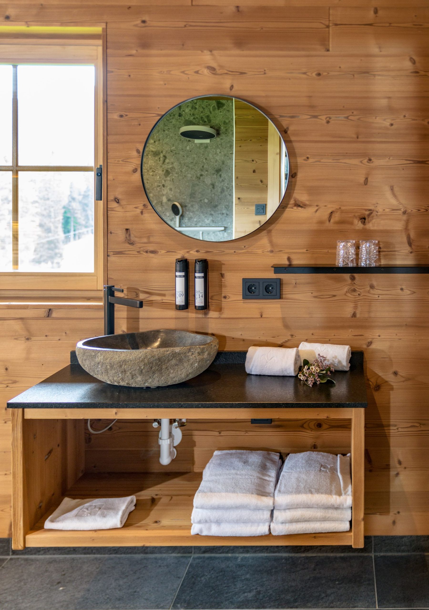 Badezimmer mit Wänden aus gebürstetem Gladiola-Holz, steinernem Waschbecken auf schwarzer Arbeitsplatte, rundem Spiegel und gefalteten Handtüchern.