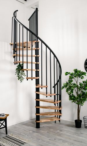 Die Spindeltreppe Berlin mit Buchenstufen und schwarzem Geländer ist an einer Galerie montiert. Der Raum ist mit Pflanzen, einer schwarzen Wanduhr, einem schwarzen Beistelltisch und einem gemusterten Teppich dekoriert.