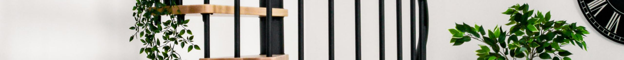 Die Spindeltreppe Berlin mit Buchenstufen und schwarzem Geländer ist an einer Galerie montiert. Der Raum ist mit Pflanzen, einer schwarzen Wanduhr, einem schwarzen Beistelltisch und einem gemusterten Teppich dekoriert.